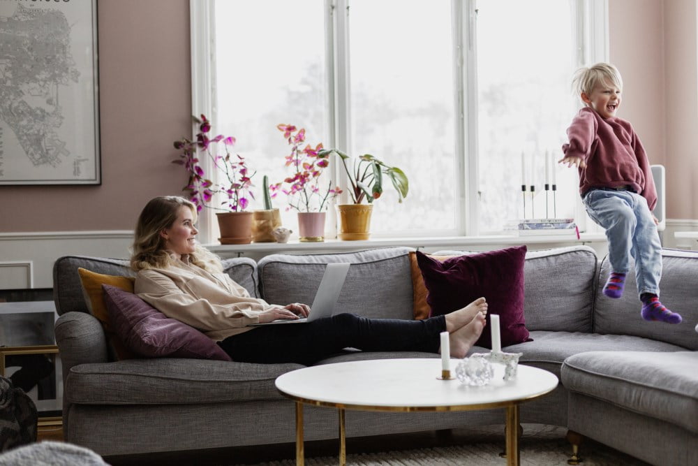 Mor som sitter i sofa og jobber, mens barnet hopper ved siden av